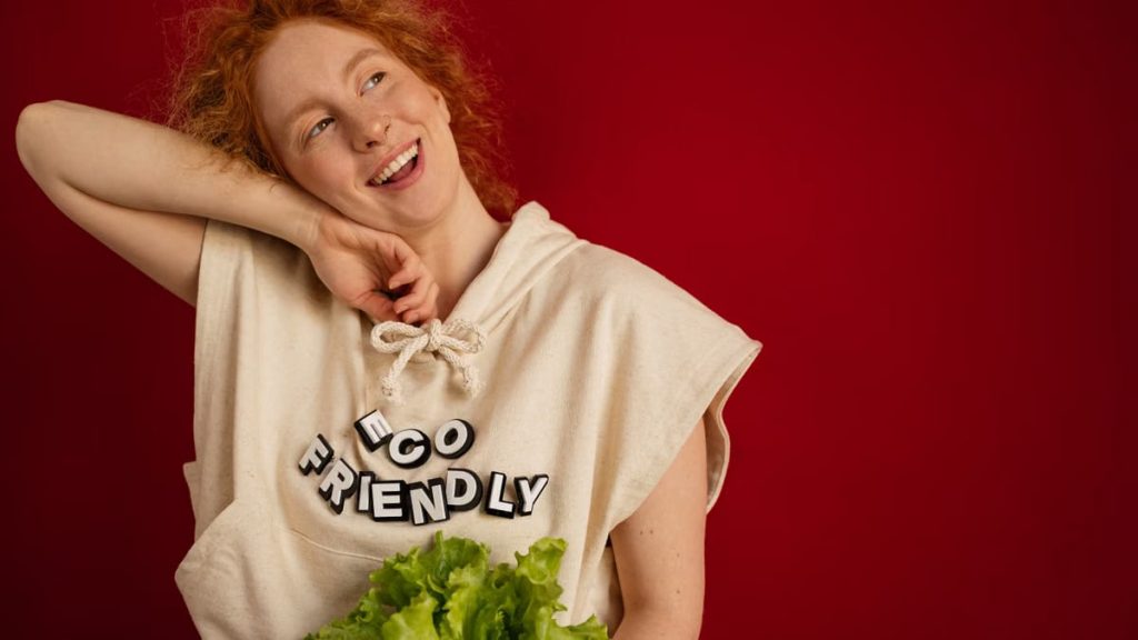 Cheerful woman with red hair in eco-friendly attire holding fresh lettuce against a vibrant red background.