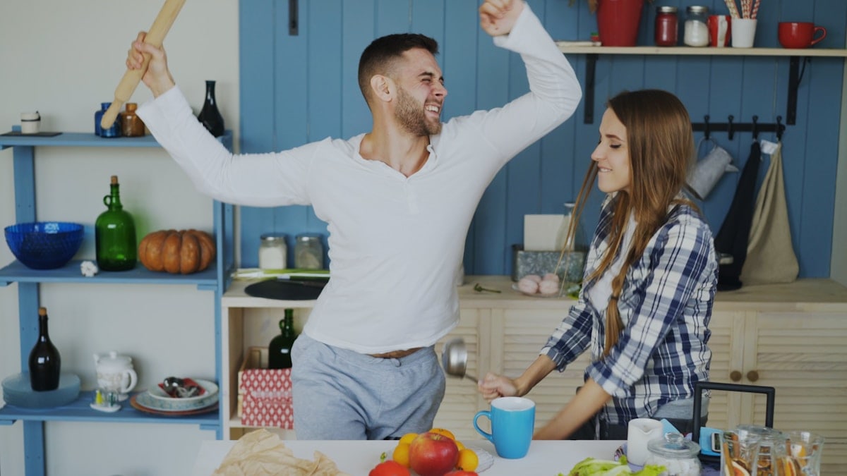 Attractive young joyful couple have fun dancing and singing while set the table for breakfast in the kitchen at home